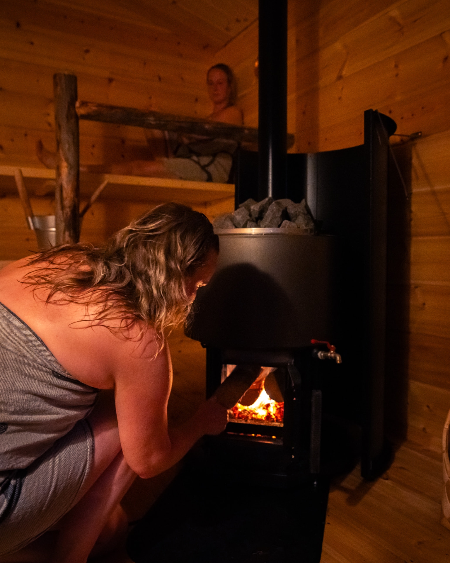 A guest wrapped in a towel feeding firewood into the glowing stove of a wood-fired sauna, warm light on her face