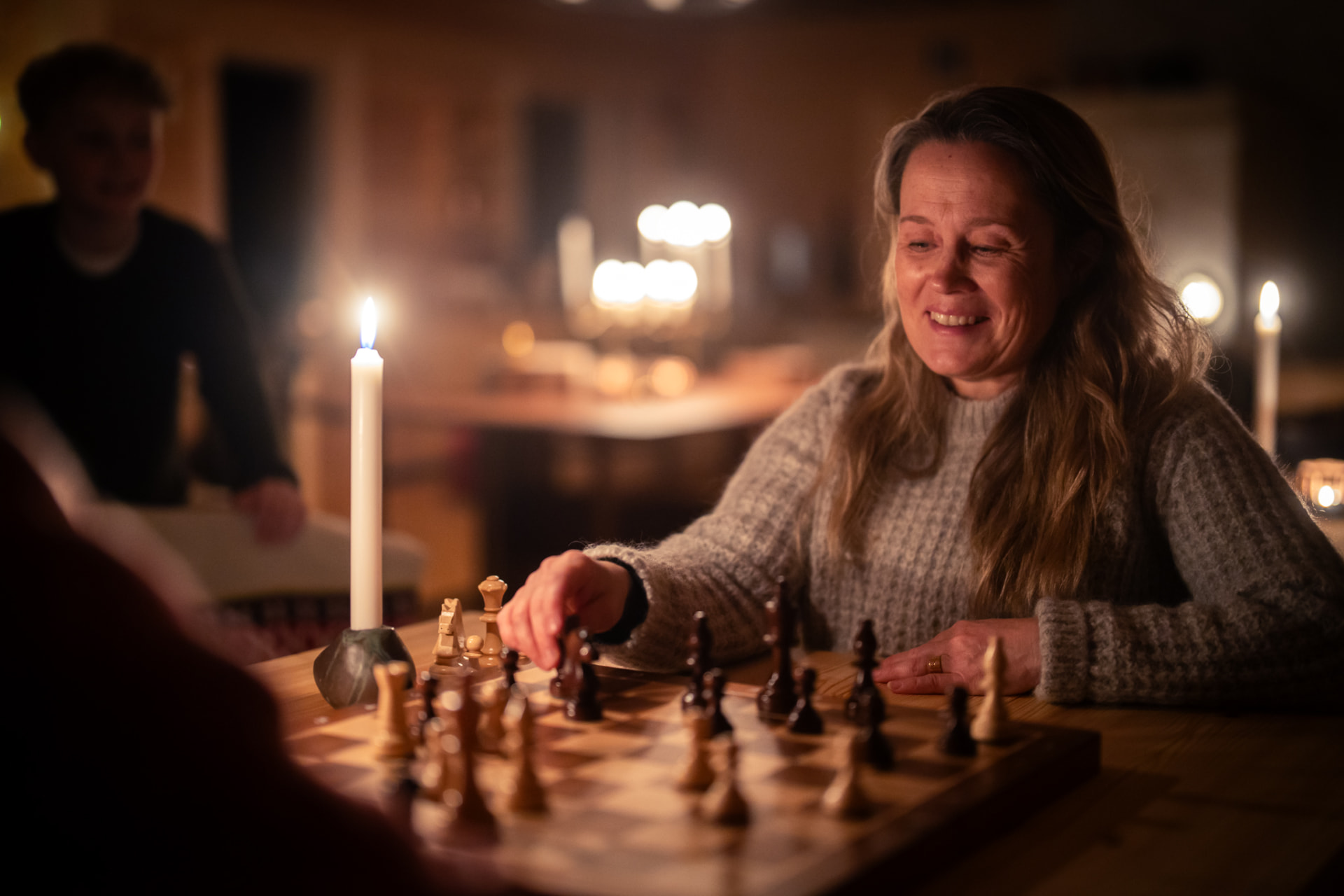 Guests playing chess at a candlelit wooden table in the Main Cabin, a child watching in the background